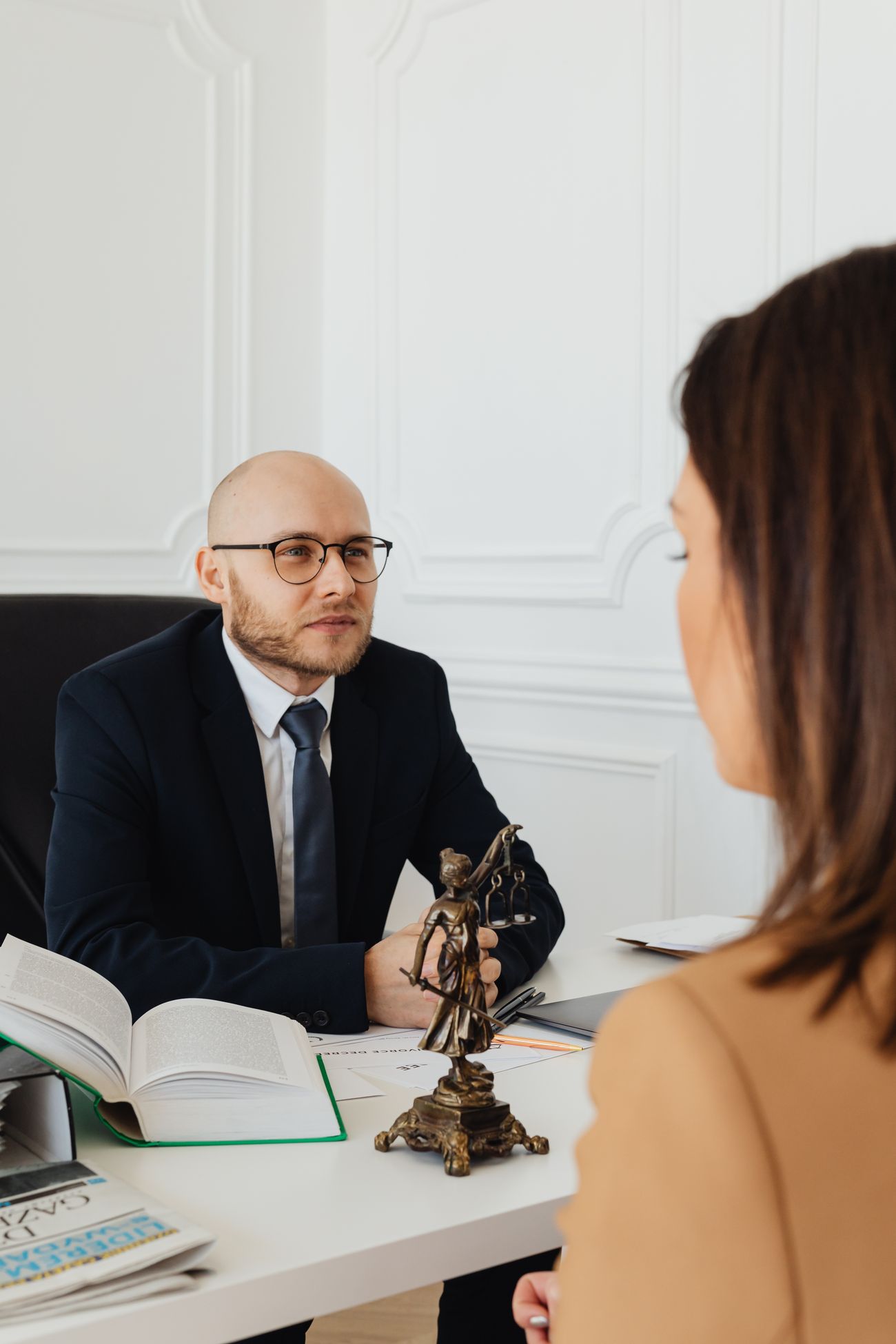 Photographie d’illustration : un avocat échange avec une cliente dans un bureau, avec des dossiers et une statuette de la Justice